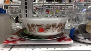 A vintage, white bowl with a brown mushroom design sitting on top of a few plates on a Goodwill shelf