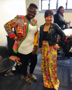 A picture of two women: the DCGF in a floral long skirt and leather jacket and another woman in a brightly patterned jacket, black pants, white shirt, and black glasses