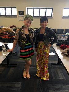 A picture of two women: the DCGF in a floral long skirt and leather jacket and a second woman in a black, green, orange, red, green, and blue striped skirt, and sleeveless jacket with patches, a black shirt, and multiple tattoos