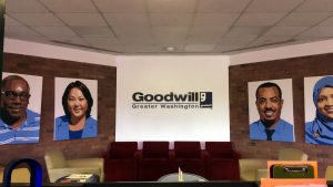 A picture of the front desk of the Goodwill of Greater Washington Headquarters. On the back wall there is a Goodwill of Greater Washington logo and for painted pictures of people who have been served by Goodwill of Greater Washington