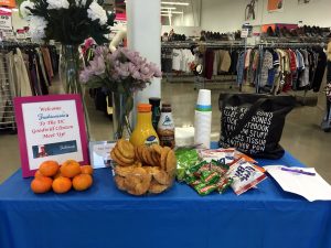 A table with a blue cloth on it at the front of the Clinton Goodwill retail store in front of a cash register with snacks like cookies, oranges, muffins, and orange juice with a sign welcoming the attendees of the MeetUp