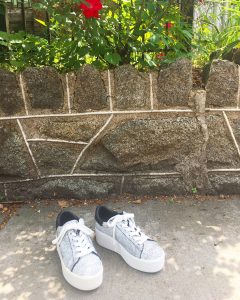 A picture of a pair of grey, platform, sneakers outside in front of a stone wall and flower bushes