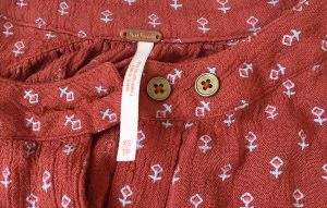 A picture of a Free People pumpkin colored blouse with gold buttons and small, white, geometric, stitch designs (A closeup of the tags which tell the blouse is extra small)