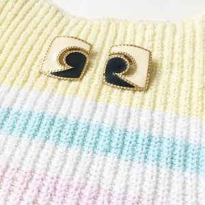 A picture of a sleeveless striped sweater that is a pale yellow, blue, pink, and white. On top of the sweater is a pair of artsy, stud, black and cream earrings with gold outlining (this picture is a close up of the earrings themselves)