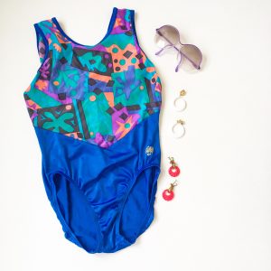 A picture of a woman's one piece bathing suit: the bottom half is blue and the top half is a combination of tropical colors in geometric shapes and palm trees. Also pictured is a pair of purple rimmed sunglasses, white circle dangle earrings, and pink circle dangle earrings