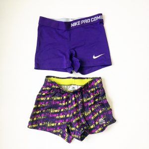 A picture of two pairs of woman's workout shorts: the pair on top are Nike ProCombat and are solid purple. the bottom are Adidas dark and light purple with a bright yellow color