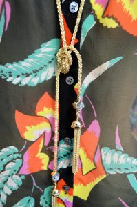 A picture of a sleeveless, black, button up, blouse with a floral patterm with a gold tassel necklace with glass beads on top of it