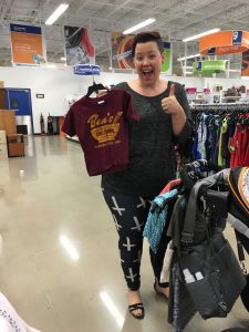 Picture of a woman at the Clinton Goodwill retail location with a cart full of items holding up a child's Ben's Chili Bowl shirt