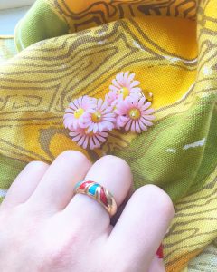 A picture of a hand wearing a multicolored gold ring, a pair of flower clip on earrings, and yellow patterned napkins