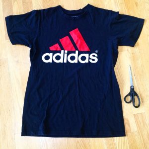 A picture of an Adidas t-shirt, it is blue with a pink three strip logo with the word adidas under the logo. There is also a pair of scissors sitting next to the shirt