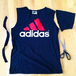 A picture of an Adidas t-shirt, it is blue with a pink three strip logo with the word adidas under the logo. There is also a pair of scissors sitting next to the shirt. The sleeves of the shirt have been cut off