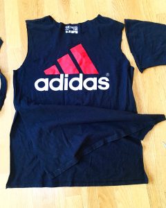 A picture of an Adidas t-shirt, it is blue with a pink three strip logo with the word adidas under the logo. There is also a pair of scissors sitting next to the shirt. The sleeves of the shirt have been cut off and the front has now been cut on both sides to create a flap in front