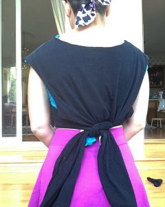 A picture of the back of a woman's midsection, she has on a purple dress, short hair, a black and white bandanna, and a DIYed tee shirt tied in a knot 