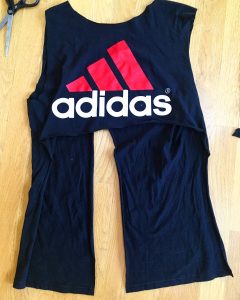 A picture of an Adidas t-shirt, it is blue with a pink three strip logo with the word adidas under the logo. There is also a pair of scissors sitting next to the shirt. The sleeves of the shirt have been cut off and the front has now been cut on both sides. The flap has now been cut and a segment has been cut out of the back to create two strips still attached to the shirt