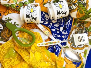 Two white coffee mugs with flowers printed on them, a green avocado cuber, a spatula and serving spoon with white handles that have floral patterns on them on top of an ornate blue table cloth and yellow patterned napkins