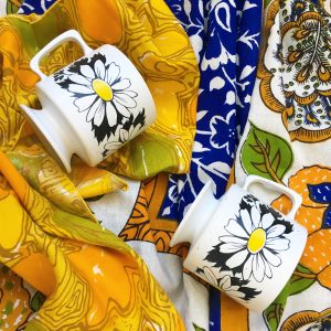 Two white coffee mugs with flowers printed on them on top of an ornate blue table cloth and yellow patterned napkins