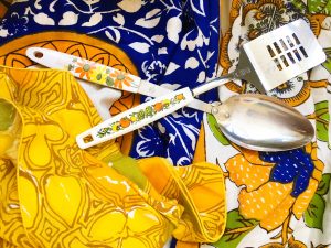 A spatula and serving spoon with white handles that have floral patterns on them on top of an ornate blue table cloth and yellow patterned napkins