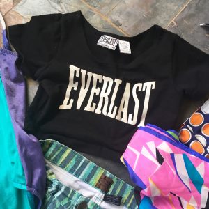A black, Everlast crop top surrounded by patterned scarves, a retro one piece bathing suit, and patterned high wasted shorts