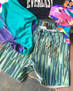 A picture of blue and green striped patterned high waisted short that have been converted from pants surrounded by a black, Everlast crop top shirt, pink patterned scarf, a retro one piece bathing suite, and gold earrings