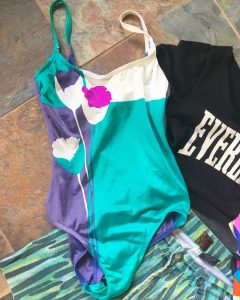 A women's, retro, one piece swim suit that is white, teal, and purple with some flower like shapes