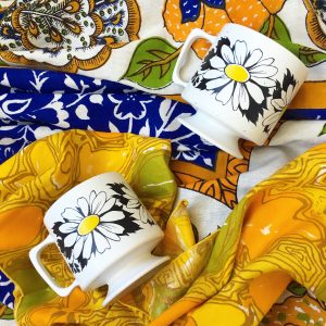 A picture of two white mugs with flowers on them. They are laying on top of a colorful table cloth