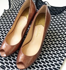 A picture of a top that is a stretchy, black and white, short sleeved, peplum blouse by Intermix in a size medium. On top of the shirt is a pair of reddish, warm gold, metallic, short, open-toed, modern, Cole Haan heels in a size 7. The picture is zoomed in on the shoes