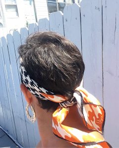 A back view of the head of a woman with short brunette hair. She is wearing gold hoop earrings and a vintage silk scarf with a black, white, and orange houndstooth pattern on it that is around her head and tied at the back