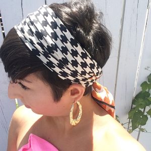 A side view of a woman with short brunette hair. She has on gold hoop earrings and a vintage silk scarf with a black, white, and orange houndstooth pattern on it that is around her head and tied at the back