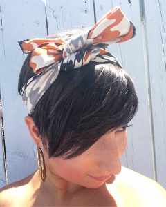 The top view of a woman's head. She has short brunette hair that covers one of her eyes and is wearing gold hoop earrings and a vintage silk scarf with a black, white, and orange houndstooth pattern on it that is around her head and tied at the top in a bow