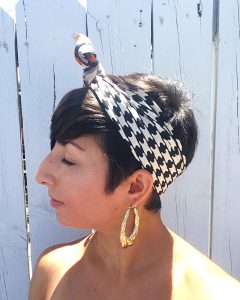 A side view of a woman with short brunette hair. She has on gold hoop earrings and a vintage silk scarf with a black, white, and orange houndstooth pattern on it that is around her head and tied at the top in a bow