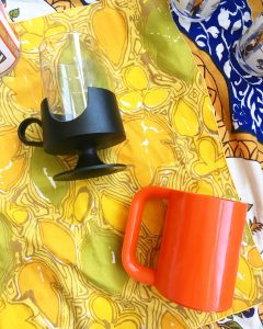 A picture of two mid-century, modern, 70's styled cups. One is all orange and blocky with a large handle and the other is glass with a black bottom and smaller handle. Both cups are laying on their sides on top of a yellow patterned table cloth