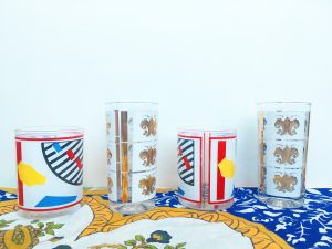 A picture of four cups sitting on top of an ornate classical table cloth. Two of the cups are short, bourbon-style, art deco, pop art cups and two of the cups are tall gold and white printed with fleur de lis on them.