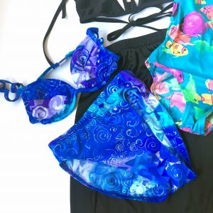 A picture of a blue, high waisted bikini with different 90's patterns on it, a one piece bathing suit that is a turquoise color with hot pink tropical fish on it, and a black bikini top and a black skirt with a pastel boarder. This particular picture focuses on the blue two piece bathing suit