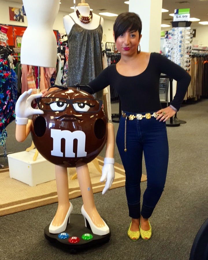 A picture of a woman in a black, long sleeved top, blue jeans, and neon shoes standing next to a large brown M&M figurine