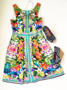 A picture of a sleeveless, bright, Maggy London dress with a tropical floral pattern on it. Next to the dress is a pair of black and white Aztec triangle patterned TOMS wedge open toed shoes. 