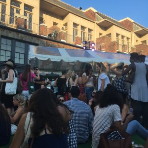 A large crowd of people in front of a covered vendor stand with buildings in the background.