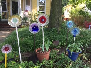 Six DIY flowers made from plates, jello molds, glass wear in different colors sitting "planted" outside of a yellow house