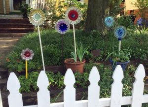 Six DIY flowers made from plates, jello molds, glass wear in different colors sitting "planted" outside of a yellow house with a white picket fence in front of them