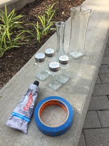 A picture of a tube of glue, blue painters tape, some clear vases, and four salt and pepper shakers sitting on a concrete wall