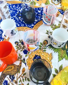 A top down view of all of the cups mentioned in this post. There are two of the art deco bourbon glasses, one of the orange block mugs, one of the glass with a black bottom and smaller handle, two of the white mugs with daisys on them, and one of the gold and white printed taller glasses with fleur de lis on them