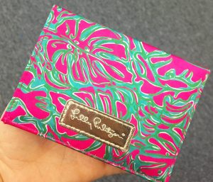 A picture of a woman holding a Lilly Pulitzer clutch purse. It has pink palm leaves against a green and aqua background. The Lilly Pulitzer logo is on a leather rectangle that is sewn to the clutch