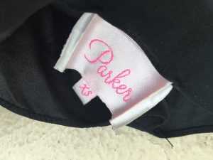 The tag of a black Parker blouse. The tag says "Parker" in pink script against a white background and "xs" in pink on a smaller tag attached to the bigger one.