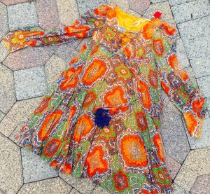 An ornately patterned 1970's Howard Hirsch dress. It has the colors yellow, orange, red, green, and purple, has a tie at the neckline and the sleeves flair at the end.