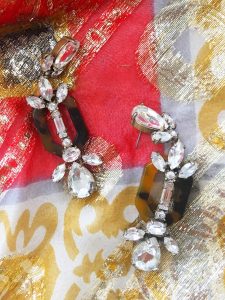 A pair of black and clear gem J Crew earrings. The gems are in different geometric shapes and the earrings dangle. They are sitting on top of a very close up picture of the gold, red, and black patterned Nelly de Grab maxi dress