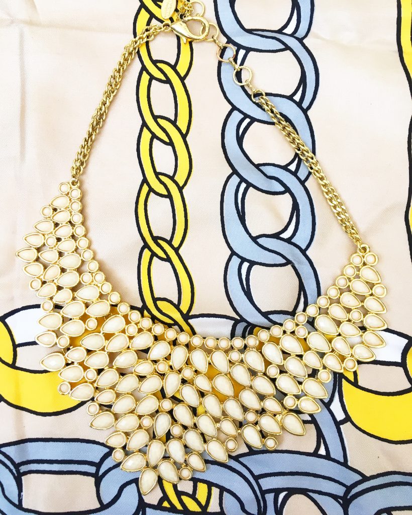 A necklace with multiple rows of cream rine stones with a gold chain. It is on top of a scarf that you can see a yellow link chain and blue link chain painted on it