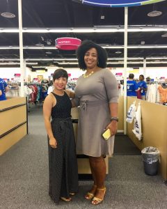 A picture of the DC Goodwill Fashionista (a short woman with short brown hair in a sleeveless black jumpsuit) and Danielle from Perfectly Good blog (a taller African American Woman in a grey dress with a yellow necklace) standing in front of the check out registers at the Centreville, VA Goodwill retail store