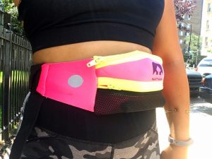 A picture of the midsection of a woman. She is wearing a black running bra, grey and black camo workout pants, and a pink Nathan fanny pack with bright yellow zippers