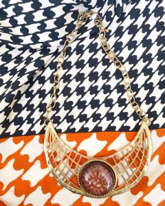 A necklace with a round, amber stone surrounded by a lattice of gold and a gold chain. This is on top of a black, orange, and white hounds tooth scarf