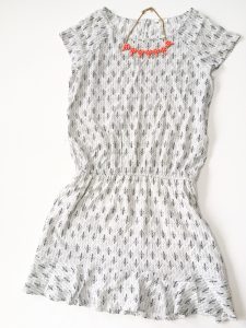 A picture of a white Joie day dress. It has a small, black, design all over it in rows that almost look like small bird's feet. The dress has very short sleeves and gets smaller around the waist. Also pictured is an orange necklace with rine stones