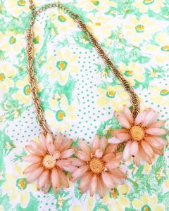 A necklace with a gold chain that connects to three pink flowers that are made of rine stones. It is on top of a light green and yellow floral scarf